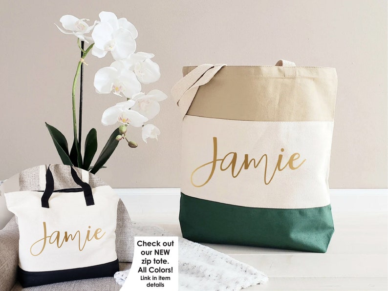 K&ouml;nnte beinhalten: Zwei personalisierte Canvas-Tragetaschen mit dem Namen "Jamie" in goldener Schrift. Die gr&ouml;&szlig;ere Tasche hat ein beige-wei&szlig;-gr&uuml;nes Farbschema. Die kleinere Tasche hat ein schwarz-wei&szlig;es Farbschema.