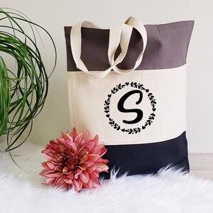 K&ouml;nnte beinhalten: Eine schwarz-wei&szlig; gestreifte Tragetasche mit grauem Oberteil. Die Tasche hat einen wei&szlig;en Canvas-Bereich mit einem schwarzen Kranz und dem Buchstaben "S" in der Mitte.