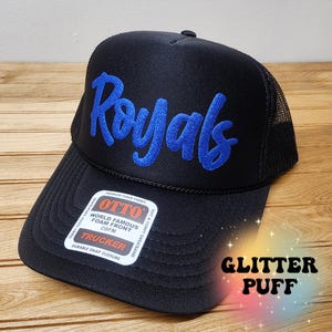 以下が含まれることがあります： 前面に青いグリッターパフで「Royals」が刺繍された黒いトラッカーハット。帽子はメッシュバックとスナップクロージャーが付いています。ラベルには「OTTO World Famous Foam Front OSFM Trucker Durable Snap Closure」と記載されています。