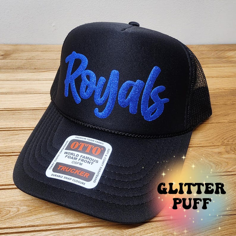 以下が含まれることがあります： 前面に青いグリッターパフで「Royals」が刺繍された黒いトラッカーハット。帽子はメッシュバックとスナップクロージャーが付いています。ラベルには「OTTO World Famous Foam Front OSFM Trucker Durable Snap Closure」と記載されています。