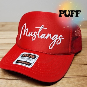 May include: Red trucker hat with a white "Mustangs" logo on the front. The hat has a mesh back and a snap closure. The hat is made by Otto World Famous Foam Front.