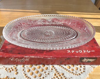 May include: Clear, oval-shaped glass serving tray with a detailed floral and geometric pattern. The tray sits on a red box with Japanese text and the name "Elizabeth". The tray's dimensions are approximately 12 inches long and 8 inches wide.