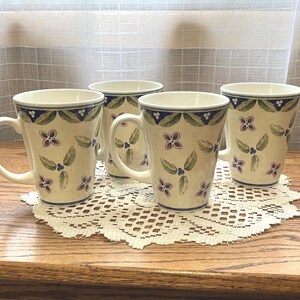 May include: A set of four cream-colored ceramic mugs featuring a hand-painted pattern of green leaves, small purple flowers, and a dark blue decorative border near the rim. Each mug has a sturdy handle and a tapered cylindrical shape.