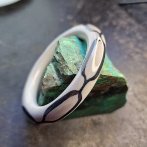 Könnte beinhalten: Ein weißes Bangel-Armband mit einem schwarzen geometrischen Muster. Das Armband liegt auf einem grünen Stein.