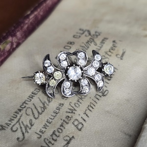 May include: A silver-toned brooch with a floral design, set with numerous clear and pale yellow gemstones. The brooch has a central flower shape with surrounding petals and two smaller gemstone clusters on either side. The brooch is displayed on a fabric surface.