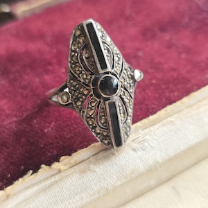 Könnte beinhalten: Ein silberfarbener Ring mit einer länglichen, schildförmigen Oberfläche. Der Ring hat einen zentralen schwarzen Stein, flankiert von zwei schwarzen vertikalen Balken und umgeben von kleineren, funkelnden Steinen. Der Ring ist vor rotem Samt platziert.