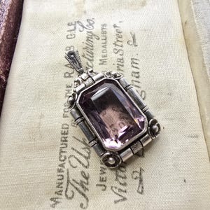 Może przedstawiać: Antyczny srebrny wisiorek z dużym, prostokątnym, jasnofioletowym kamieniem szlachetnym. Wisiorek ma ozdobne srebrne detale wokół kamienia i dekoracyjne krawędzie. Widoczny jest tekst "MANUFACTURED FOR THE RAB GLE".