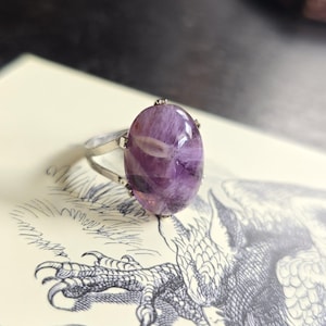 Könnte beinhalten: Ein Silberring mit einem großen, ovalen Amethyst-Edelstein. Der Amethyst zeigt verschiedene lila Farbtöne und interne Muster. Die Fassung des Rings ist schlicht und betont den Edelstein. Der Ring liegt auf einer Seite mit einer detaillierten Illustration.