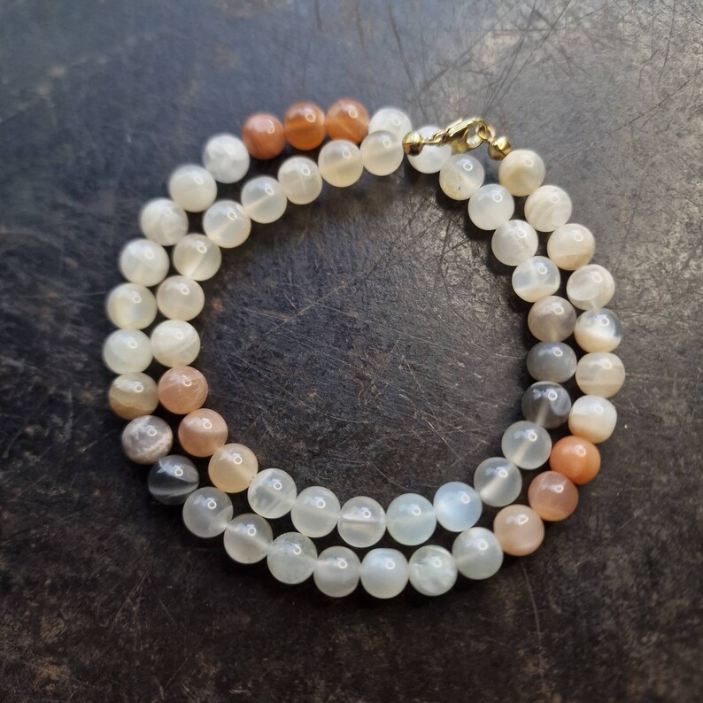 May include: A necklace made of round, white, gray, and orange moonstone beads. The necklace has a gold clasp.