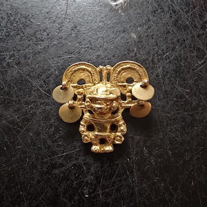 Könnte beinhalten: Eine goldfarbene, verzierte Brosche mit einer zentralen Figur und kreisförmigen Anhängern. Die Brosche hat ein symmetrisches Design mit detaillierten Schnitzereien und einer polierten Oberfläche. Der Hintergrund ist eine dunkle, strukturierte Oberfläche.