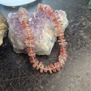 May include: A pink and clear quartz gemstone bracelet with gold spacers. The bracelet is made of flat, oval-shaped beads.