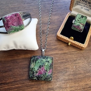 Op de afbeelding: Een set van drie sieraden met een groene en roze edelsteen. De set bevat een armband, een hanger en een ring. De sieraden worden gepresenteerd op een houten oppervlak.