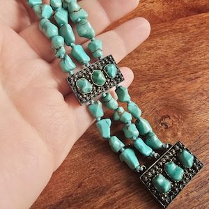 May include: A turquoise beaded necklace with three strands. The necklace features rectangular metal accents with turquoise stones. The beads are a light blue-green color. The necklace is held in a hand.
