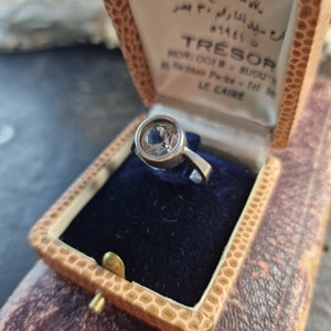 Puede incluir: Un anillo de plata con una piedra redonda transparente en una caja de anillo de terciopelo azul. La caja es de cuero marrón con un cierre dorado. La caja tiene el texto "TRÉSOR HORLOGER-BIJOUTERIE 55, Gamal Pacha - Tel 569425 LE CAIRE" impreso en el interior.
