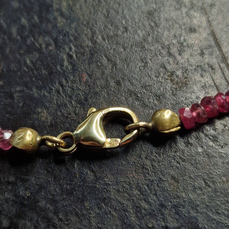 May include: Close-up of a gold clasp on a pink gemstone necklace. The clasp is a lobster claw style with a small, round bead on the end.