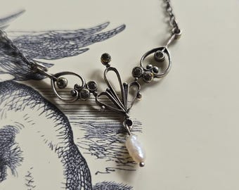 May include: A silver-toned necklace with an ornate, symmetrical design. The necklace features a central floral-like element with small, round embellishments and a teardrop-shaped pearl pendant. The chain is delicate, and the necklace is set against a vintage-style background.