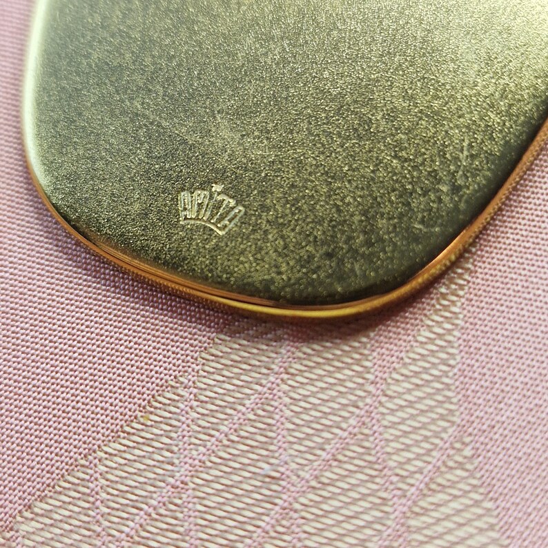 May include: Close-up of a gold-coloured, oval-shaped compact mirror with the word "APITA" engraved in a crown design.