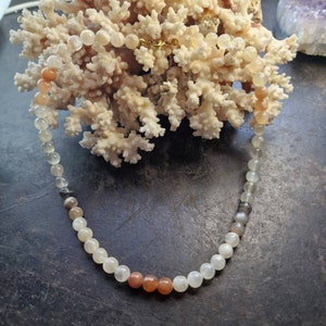 May include: A white, gray, and orange gemstone necklace with a gold clasp. The necklace is made of round beads and is displayed on a piece of coral.