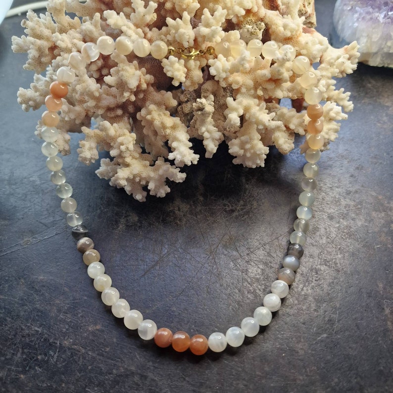 May include: A white, gray, and orange gemstone necklace with a gold clasp. The necklace is made of round beads and is displayed on a piece of coral.