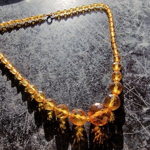 May include: An amber-coloured vintage necklace with faceted beads of different sizes. The necklace has a delicate chain and a clasp. The beads have a warm, golden hue, reflecting light. The necklace is displayed on a dark, textured surface.