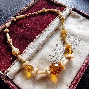 May include: A vintage necklace made of cream-coloured and amber glass beads. The necklace is displayed in a red velvet-lined box with a white satin lining. The box has a label that reads "The Us Bufacturen JEWELLF Victor Works Vittoria Street, Birmingham. ERSMITHS. MEDALLIST"