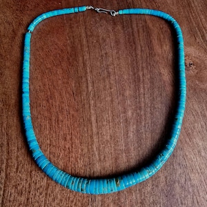 Peut inclure: Un collier de perles turquoise avec un fermoir argenté. Le collier est composé de petites perles rondes et plates de différentes nuances de bleu-vert. Les perles sont enfilées pour former une forme circulaire. Le collier est présenté sur une surface en bois.