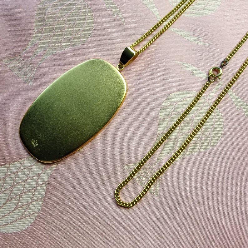 May include: A gold chain necklace with a large oval pendant. The pendant is smooth and has a small crown marking on the back.