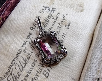 May include: A silver pendant with a clear, faceted gemstone with pink and purple hues. The pendant is in an ornate, Art Deco style setting. The pendant is on a white fabric background with text that reads "Manufactured for the Rass Gle The Usher M Jewellers, Silver Medallists Vittoria Waring Co. 81, St. Street, Birmingham."