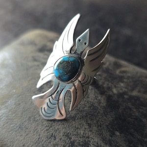 Op de afbeelding: Een zilveren ring met een turquoise steen in het midden, gevormd als een gestileerde vogel met uitgespreide vleugels.