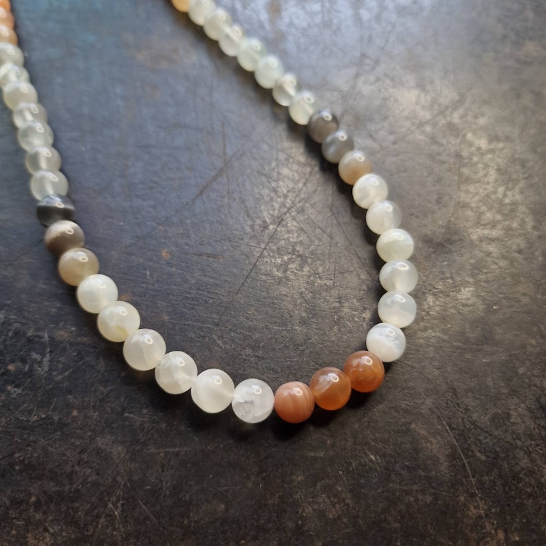 May include: A white and orange gemstone beaded necklace. The beads are round and have a smooth, polished finish. The necklace is made with a variety of stones, including white moonstone, gray moonstone, and orange sunstone.