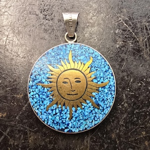 May include: A circular pendant with a silver loop. It features a gold sun design set against a blue mosaic background. The sun has a smiling face and radiating rays. The pendant is likely made of metal and stone.