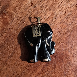 May include: A black elephant pendant with gold accents. The pendant features a detailed carving of an elephant with gold-colored tusks, feet, and a decorative element on its back. The pendant is suspended from a gold-colored bail.