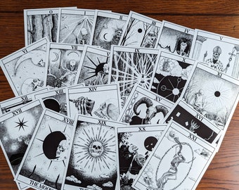 Op de afbeelding: Een set van 22 zwart-witte tarotkaarten met een schedelmotief. De kaarten liggen in een cirkel op een houten oppervlak. De kaarten zijn genummerd van 0 tot 21 en bevatten de grote arcana-kaarten: De Dwaas, De Tovenaar, De Hogepriesteres, De Keizerin, De Keizer, De Paus, De Geliefden, De Wagen, Kracht, De Kluizenaar, Het Rad van Fortuin, Rechtvaardigheid, De Gehangene, De Dood, Matigheid, De Duivel, De Toren, De Ster, De Maan, De Zon, Het Oordeel en De Wereld.