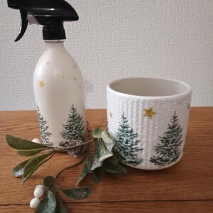 Op de afbeelding: Witte spuitfles en keramische pot met een kerstboomontwerp. De fles heeft een zwarte sproeikop en gele steraccenten. De pot heeft een getextureerd oppervlak en vergelijkbare boom- en sterontwerpen. Een takje groen met witte bessen ligt ervoor.