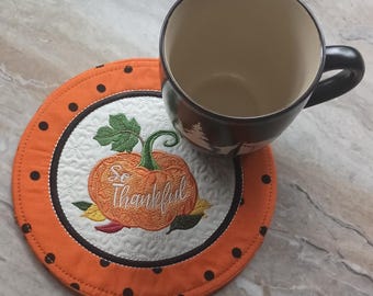 May include: An orange and white quilted pumpkin coaster with the words "So Thankful" embroidered on it. A black and brown mug with a forest scene sits on the coaster. The coaster has black polka dots around the edge.