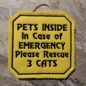 Bright yellow felt sign embroidered with PETS INSIDE In case of EMERGENCY Please Rescue 3 CATS in bold letters with black thread. 
Has yellow felt on the back with paracord for hanging. Edges are done with pinking shears. Size approx. 5x5 square