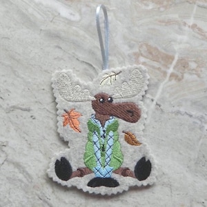 May include: A felt moose ornament with embroidered details. The moose has large antlers, a brown snout, and is wearing a green and blue sweater. Autumn leaves are scattered around the moose. A silver ribbon is attached for hanging.