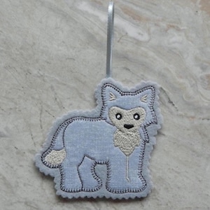 May include: A light blue felt wolf ornament with embroidered details. The wolf has a white face, beige chest, and a gray stitched outline. A silver ribbon is attached for hanging. The ornament is designed to be a decorative item.