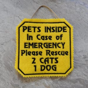 May include: A yellow felt sign with scalloped edges and a hanging string. The sign features black embroidered text that reads: PETS INSIDE In Case of EMERGENCY Please Rescue 2 CATS 1 DOG. The sign has a black border around the text.