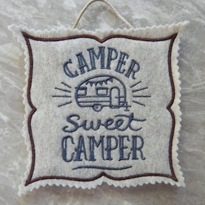 May include: A cream-colored felt wall hanging with the words "CAMPER Sweet CAMPER" embroidered in blue. The design includes a camper illustration and a brown scalloped border. A beige string is attached for hanging.
