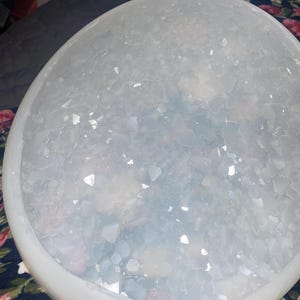 May include: A white, round, shallow bowl filled with small, clear, iridescent crystals. The bowl is made of a smooth, white material and has a slightly raised rim.