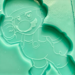 May include: A green silicone mould with a raised design of a cartoon character. The character is wearing a hat with an "M" on it and has a moustache.