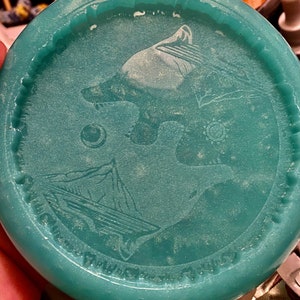 Op de afbeelding: Een tealblauwe schijf met een wit ontwerp van een wolf die huilt naar de maan. De schijf heeft een getextureerd oppervlak en een verhoogde rand.