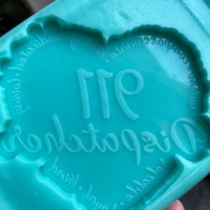 May include: A teal blue silicone mold shaped like a heart with the words "Origami" and "911" inside. The mold is for making resin crafts.