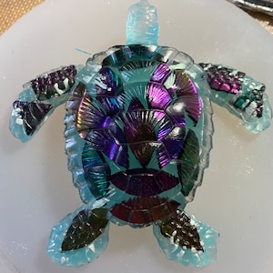 Puede incluir: Una tortuga de resina azul turquesa con detalles iridiscentes morados y verdes. El caparazón de la tortuga está hecho de piezas individuales de resina que están dispuestas en un patrón de mosaico.