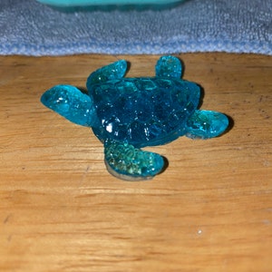 Pode incluir: Uma pequena figura de tartaruga marinha de vidro azul. A tartaruga está deitada de costas com as pernas e a cabeça estendidas.