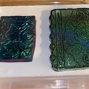 May include: Two green and iridescent resin book-shaped ornaments with glitter accents. The book on the left has a more textured surface, while the book on the right has a smooth surface with glitter embedded in the resin.