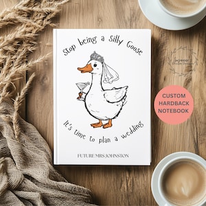 Può includere: Un taccuino con copertina rigida bianca con un'oca dei cartoni animati con un velo e una tiara, che tiene in mano un cocktail. Il testo recita "Stop being a Silly Goose, It's time to plan a wedding, Future Mrs. Johnston." Un cerchio rosa dice "Custom Hardback Notebook."