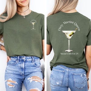 Pode incluir: Uma camiseta verde com o texto "Dirty Martini Queen" e uma taça de martini com azeitonas nas costas. A frente da camiseta tem um gráfico de taça de martini menor e o texto "Social Club Est 24".