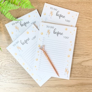 May include: Four white notepads with the text "TO DO before I DO" and champagne glass illustrations. A rose gold pen with a diamond-shaped top rests on one notepad. The pads have lined pages and heart-shaped check boxes.